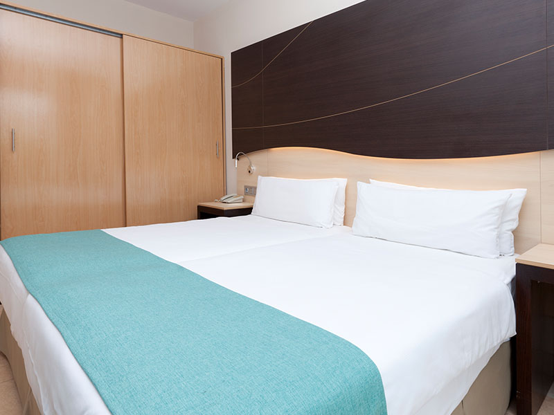 Hotel room at Impressive Playa Granada with two single beds, teal bed runners, and minimalist wood and dark panel decor.