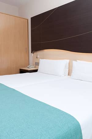 Hotel room at Impressive Playa Granada with two single beds, teal bed runners, and minimalist wood and dark panel decor.