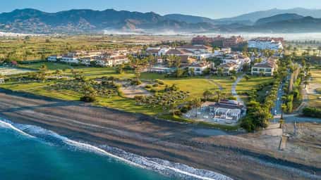 Aerial view showcasing Hotel Impressive's beachfront location, expansive lawns, sandy beach, and surrounding mountains.
