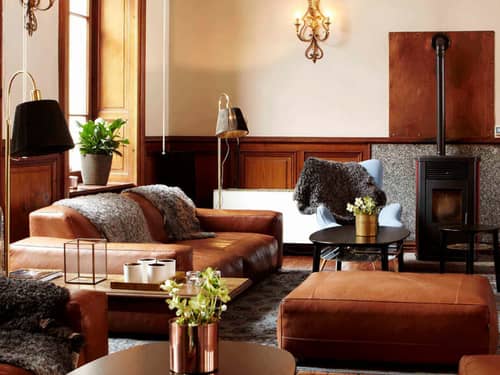 Luxurious hotel lounge with leather sofas, armchairs, plants, vintage lighting, and a bookshelf in a wood-finished interior.