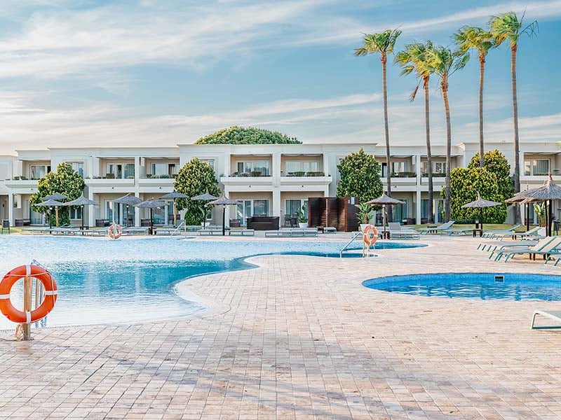 View of Hotel Vincci Costa Golf with a large swimming pool, sun loungers, palm trees, and modern hotel buildings in the background.