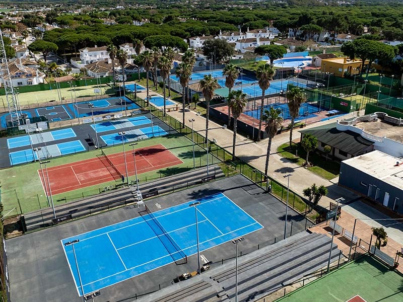 Aerial view of Hotel Vincci Costa Golf's tennis courts, featuring blue and red surfaces amid lush green landscape and nearby homes.