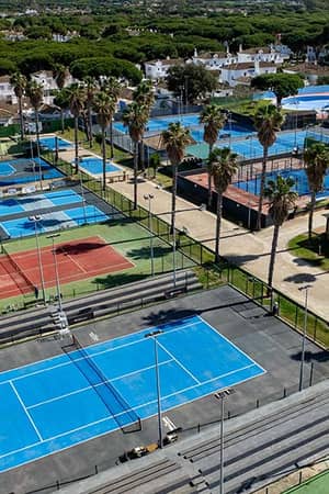 Aerial view of Hotel Vincci Costa Golf's tennis courts, featuring blue and red surfaces amid lush green landscape and nearby homes.