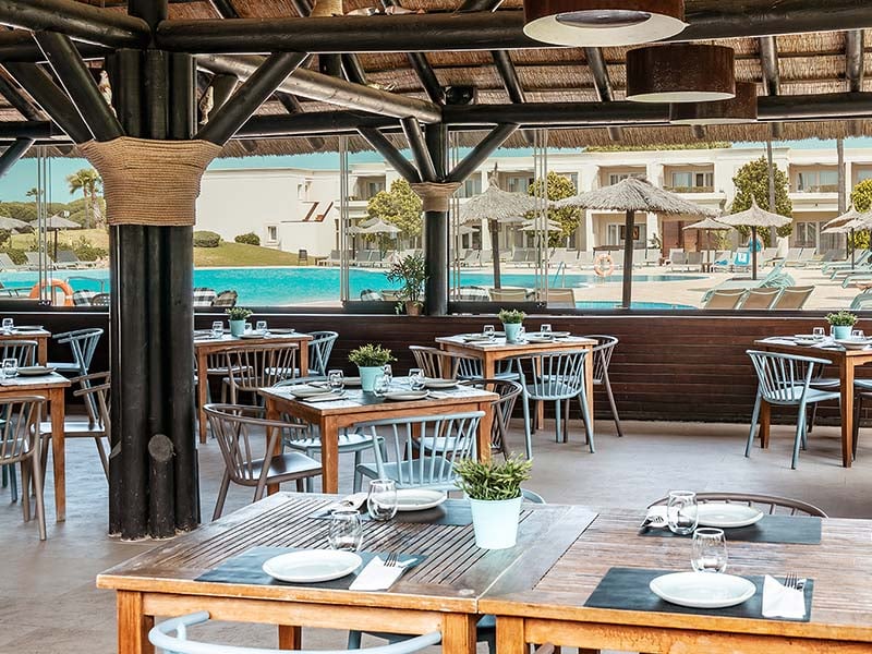 Outdoor dining area at Hotel Vincci Costa Golf with wooden tables, a view of the pool, sun loungers, and the hotel in the background.