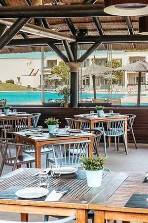 Outdoor dining area at Hotel Vincci Costa Golf with wooden tables, a view of the pool, sun loungers, and the hotel in the background.
