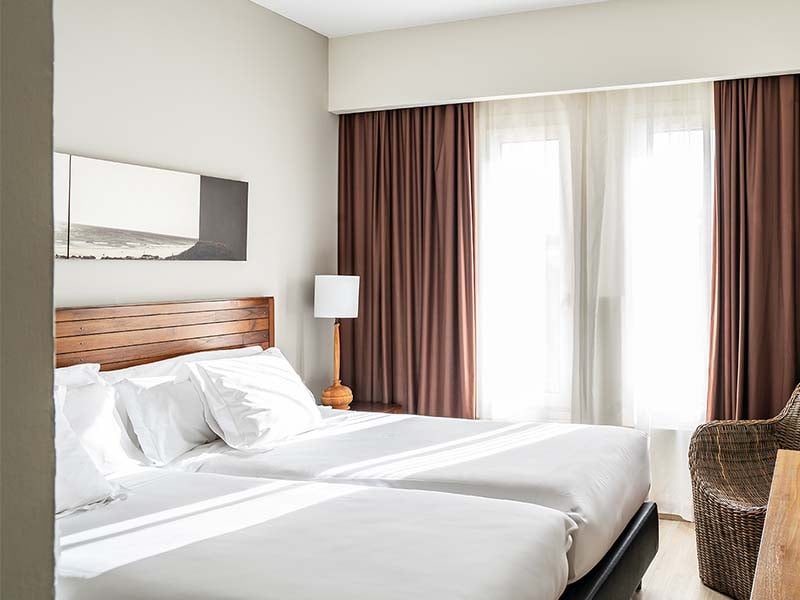 Hotel room at Vincci Costa Golf with twin beds, white bedding, wooden headboard, wicker chair, and brown curtains.