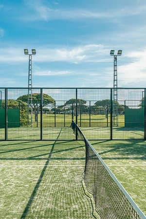 Tennis court at Hotel Vincci Costa Golf with green turf, wire fences, floodlights, and palm trees in a blue sky backdrop.