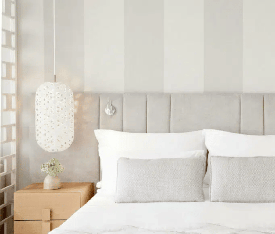 Interior of Ikos Aria Deluxe Junior Suite with a king-sized bed, grey and white pillows, pendant lamp, and bedside table with vase.