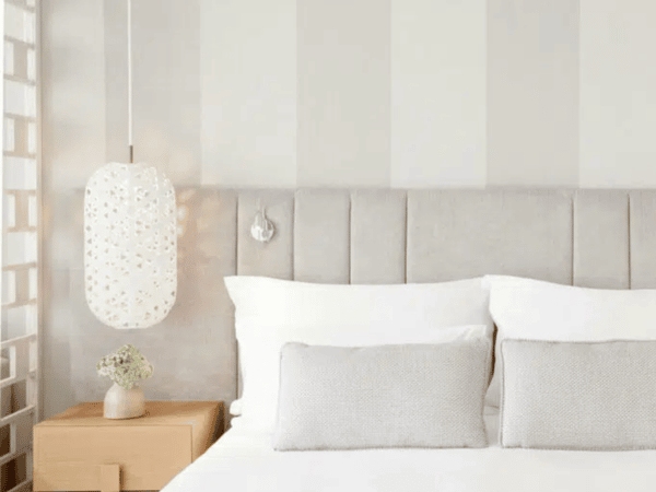 Interior of Ikos Aria Deluxe Junior Suite with a king-sized bed, grey and white pillows, pendant lamp, and bedside table with vase.