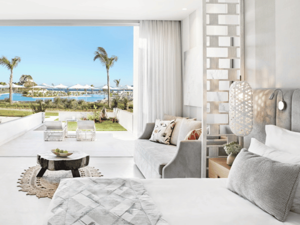 Ikos Aria Deluxe Junior Suite with elegant interior, private garden, and pool view, featuring a plush bed, seating area, and sun loungers.