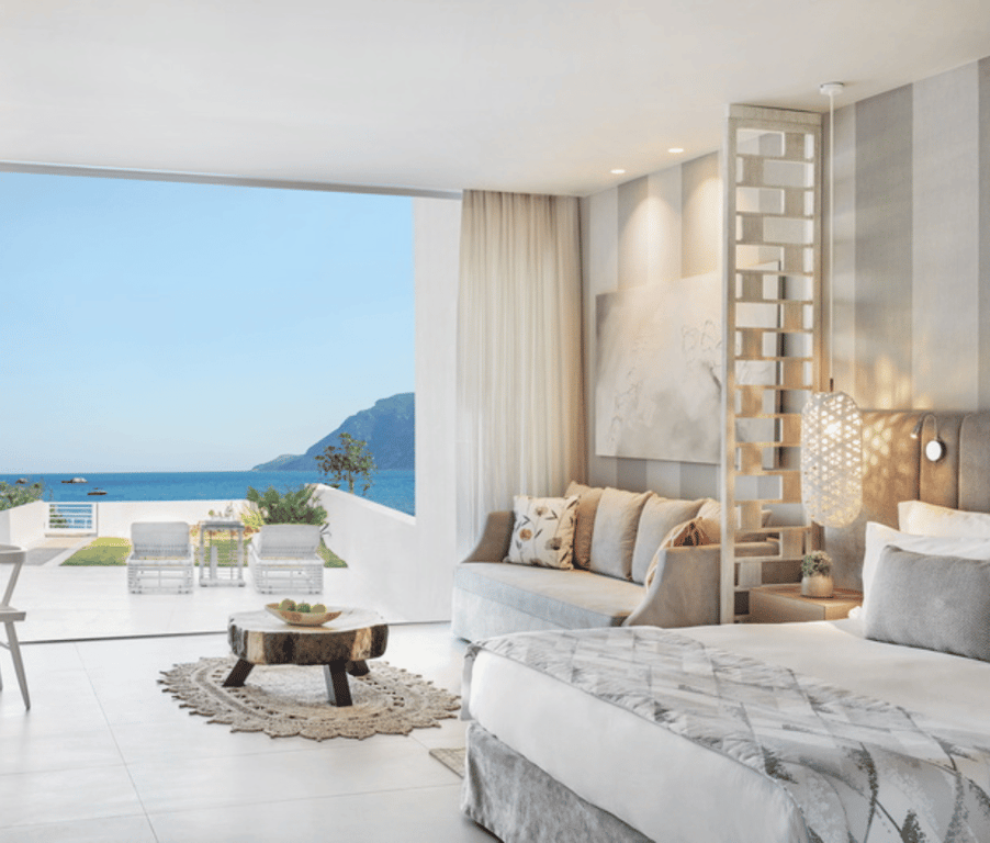 Ikos Aria deluxe junior suite with private garden and sea view, featuring an open-plan room with modern decor, a double bed, seating area, and a terrace overlooking the sea.