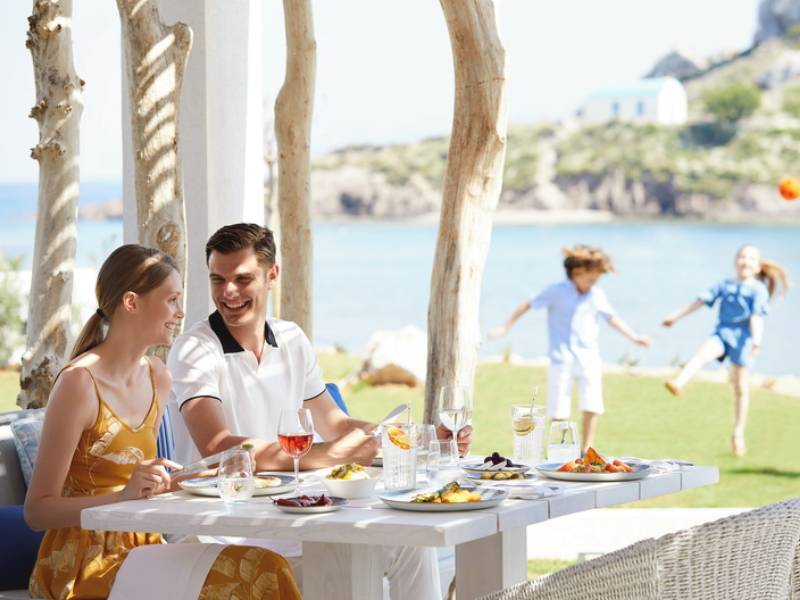 Couple dining outdoors at Ikos Aria with a variety of dishes, while children play on the lawn, overlooking the sea.