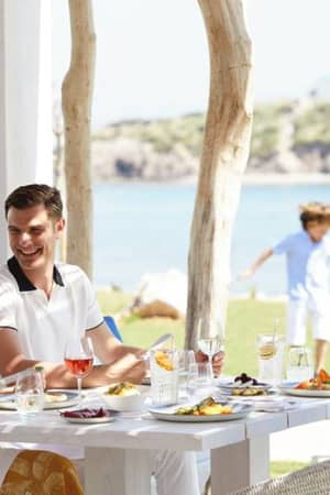 Couple dining outdoors at Ikos Aria with a variety of dishes, while children play on the lawn, overlooking the sea.