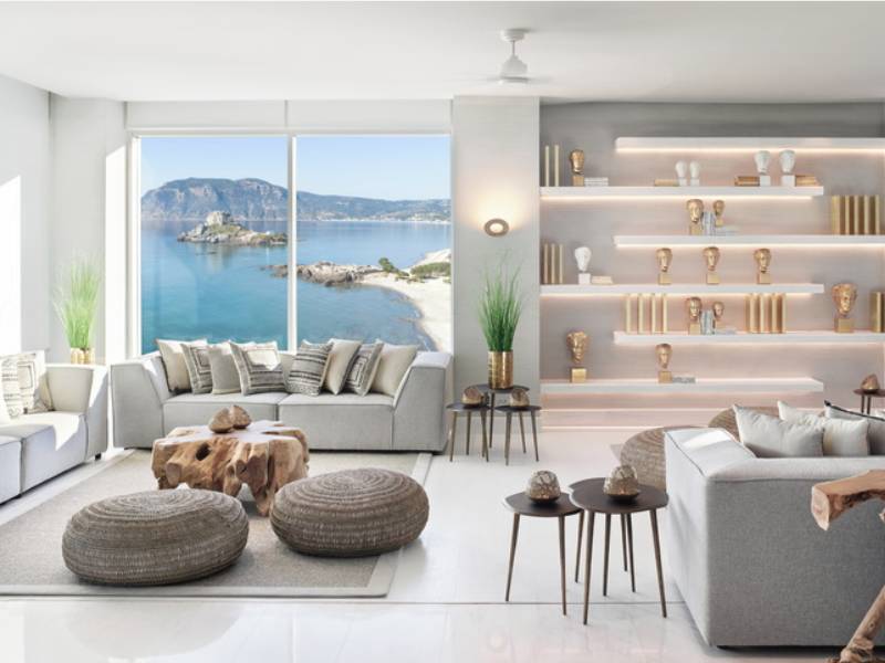 Ikos Aria hotel lobby with modern decor, neutral seating, sea view through large windows, wooden tables, and illuminated decorative shelving.