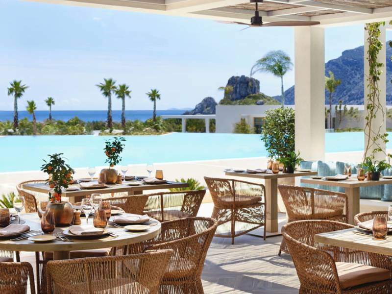 Outdoor dining tables set with crockery at Ikos Aria, with views of an infinity pool, palm trees, and the sea.