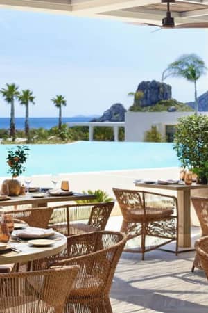 Outdoor dining tables set with crockery at Ikos Aria, with views of an infinity pool, palm trees, and the sea.