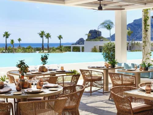 Outdoor dining tables set with crockery at Ikos Aria, with views of an infinity pool, palm trees, and the sea.