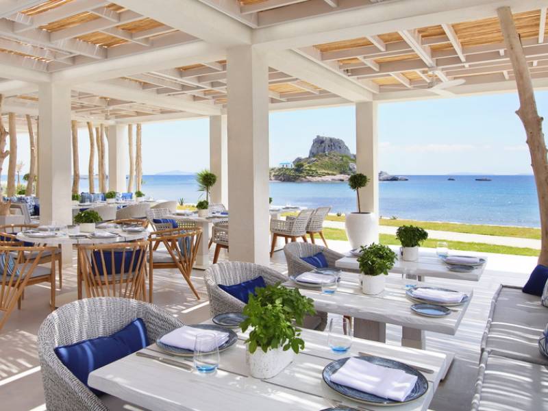 Outdoor dining area at Ikos Aria with white tables, blue accents, rattan chairs, and a sea view.
