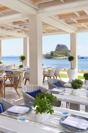 Outdoor dining area at Ikos Aria with white tables, blue accents, rattan chairs, and a sea view.
