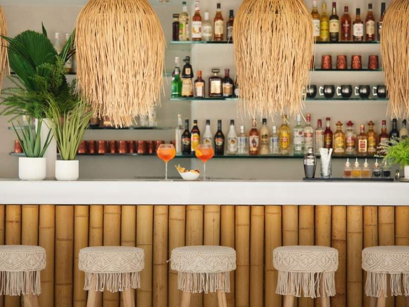 Ikos Aria Gallery bar with bamboo counter, woven stools, rattan pendant lights, and spirits on glass shelves, with two orange cocktails on the counter.