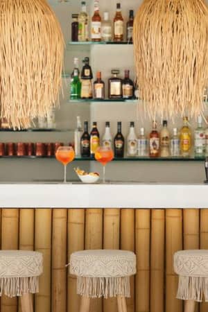 Ikos Aria Gallery bar with bamboo counter, woven stools, rattan pendant lights, and spirits on glass shelves, with two orange cocktails on the counter.