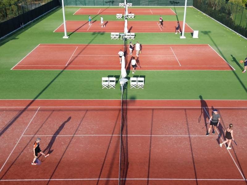 Overhead view of tennis courts at Ikos Aria with players and sunlight casting shadows, surrounded by greenery.
