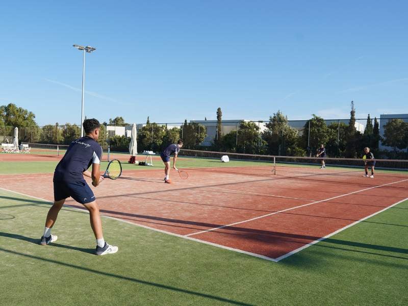 Tennis doubles match in progress on a green and red clay court at Ikos Aria, with players actively engaged under sunny skies.