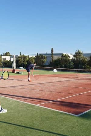 Tennis doubles match in progress on a green and red clay court at Ikos Aria, with players actively engaged under sunny skies.