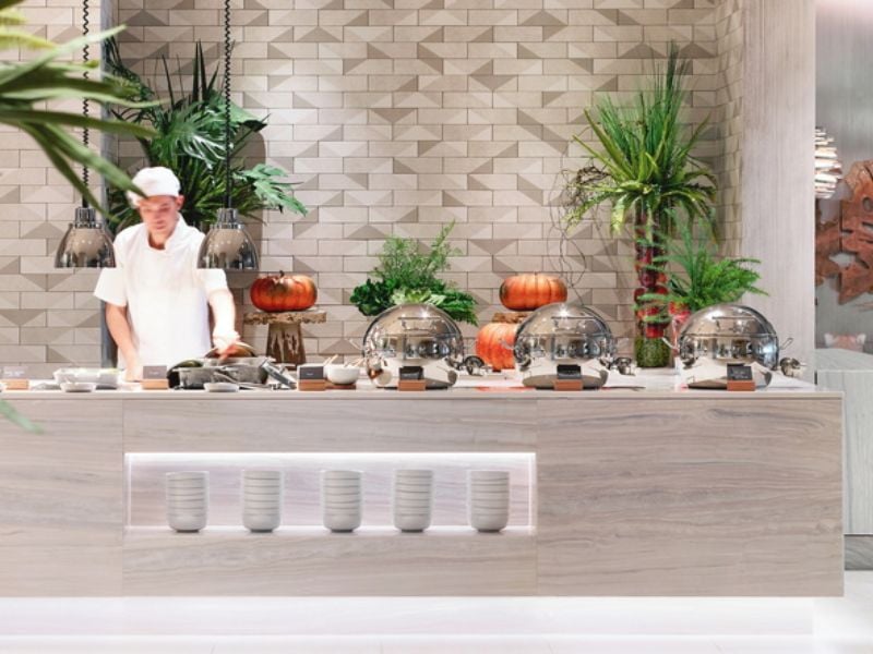 Ikos Aria dining area with a chef at a buffet table, featuring pumpkins, silver cloches, and green plants in a modern setting.