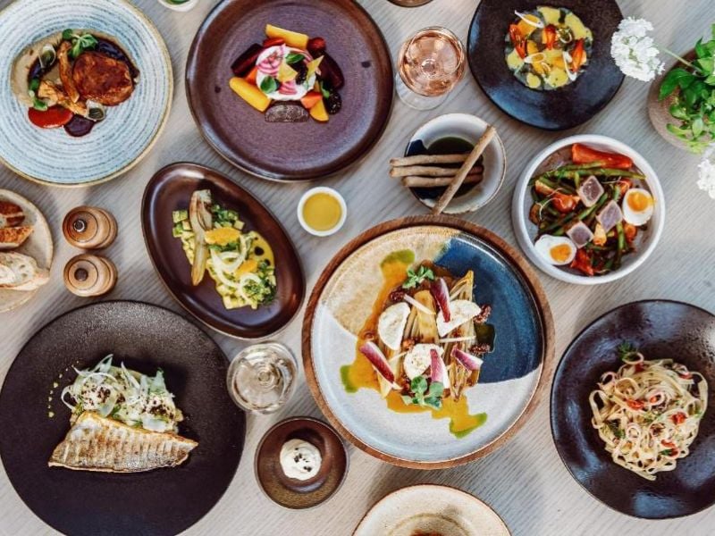 Overhead view of gourmet dishes at Ikos Aria, displaying mixed foods such as grilled fish, pasta, salads, and desserts on elegant plates.