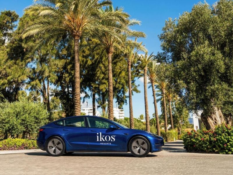 Tesla car with Ikos Resorts logo parked on a road lined with palm trees and greenery.