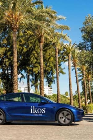 Tesla car with Ikos Resorts logo parked on a road lined with palm trees and greenery.