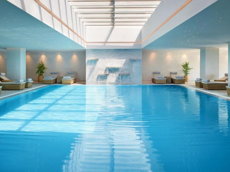 Luxurious indoor swimming pool at Ikos Aria with skylight, surrounded by sun loungers and elegant decor.