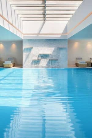 Luxurious indoor swimming pool at Ikos Aria with skylight, surrounded by sun loungers and elegant decor.