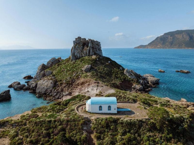 Aerial view of a small white chapel with blue roof on a rocky hill near the Aegean Sea, with cliffs and hills in the background.