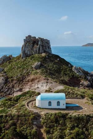Aerial view of a small white chapel with blue roof on a rocky hill near the Aegean Sea, with cliffs and hills in the background.