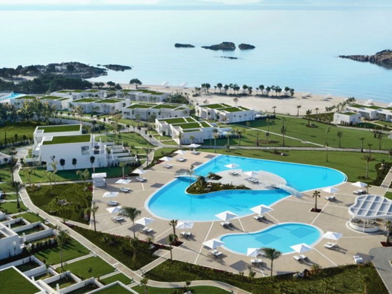 Aerial view of Ikos Aria resort, featuring a large swimming pool, white buildings with green roofs, and proximity to a sandy beach and ocean.