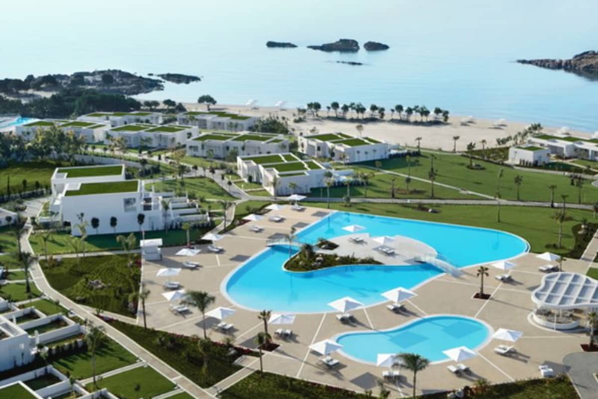 Aerial view of Ikos Aria resort, featuring a large swimming pool, white buildings with green roofs, and proximity to a sandy beach and ocean.
