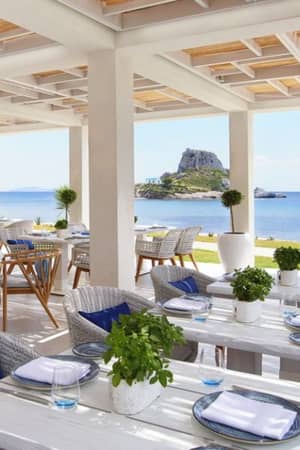 Outdoor dining area at Ikos Aria with tables, greenery, and a view of the coast and a rocky islet with a white chapel.