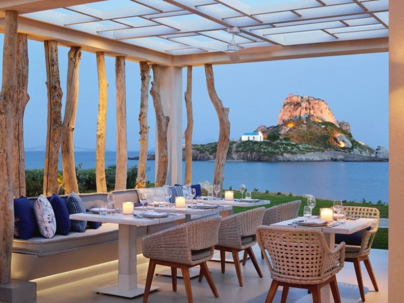 Outdoor dining area at Ikos Aria with candle-lit tables and wicker chairs, overlooking a scenic island with a church on a rocky hill.