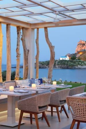 Outdoor dining area at Ikos Aria with candle-lit tables and wicker chairs, overlooking a scenic island with a church on a rocky hill.