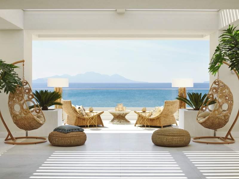 Veranda at Ikos Aria with hanging chairs, wicker armchairs, and a coffee table overlooking the sea and distant mountains.