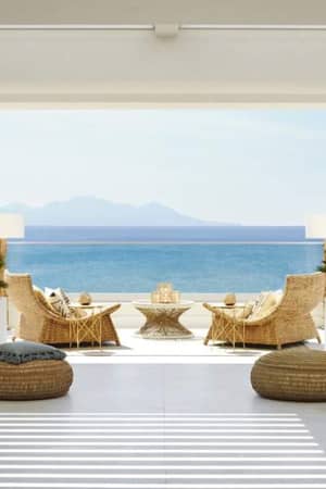 Veranda at Ikos Aria with hanging chairs, wicker armchairs, and a coffee table overlooking the sea and distant mountains.