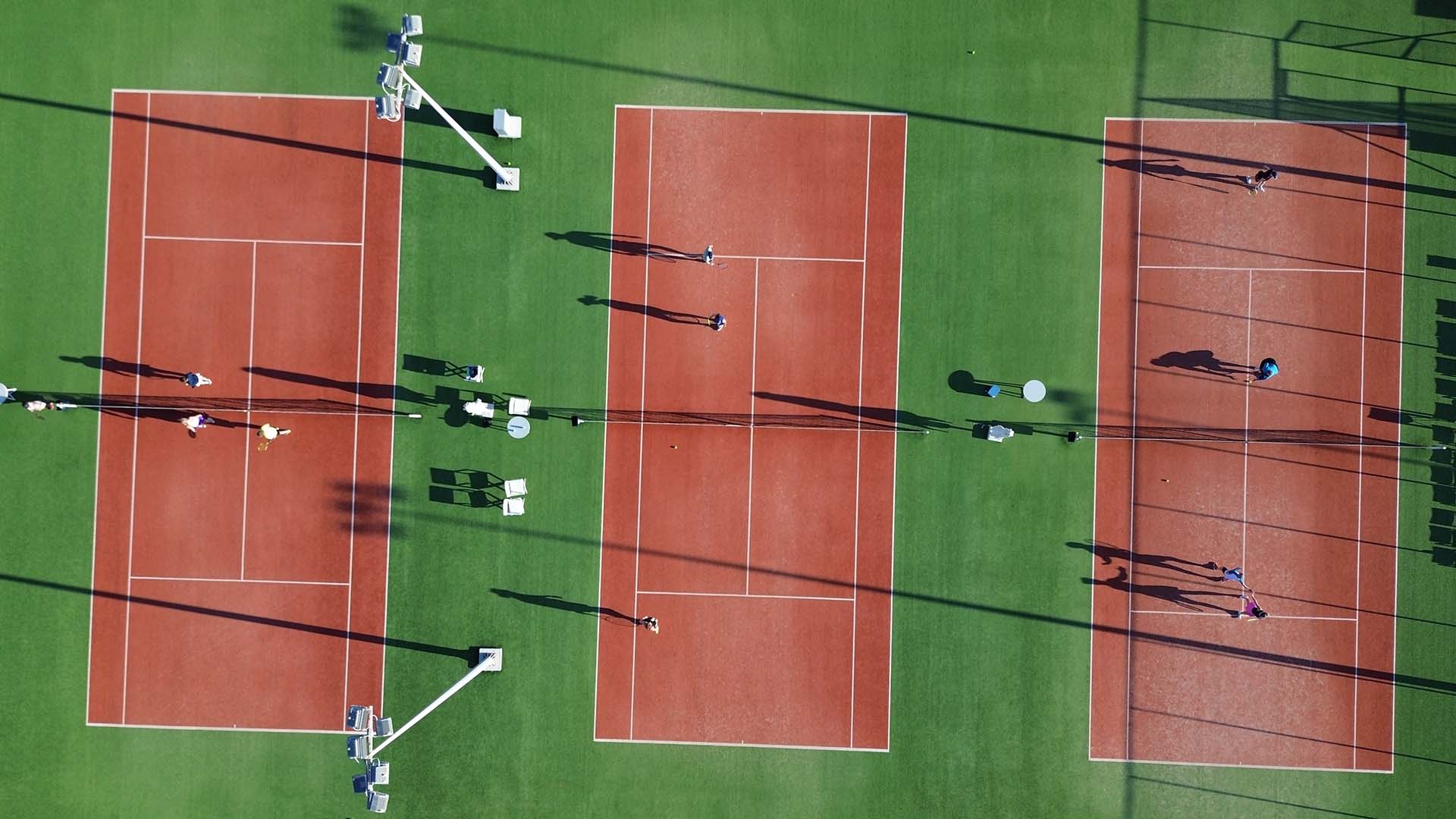 Aerial view of three clay tennis courts at Ikos Aria with players visibly engaged and shadows cast in the sunlight.