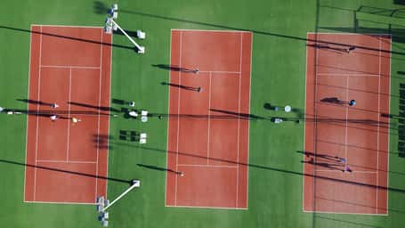 Aerial view of three clay tennis courts at Ikos Aria with players visibly engaged and shadows cast in the sunlight.