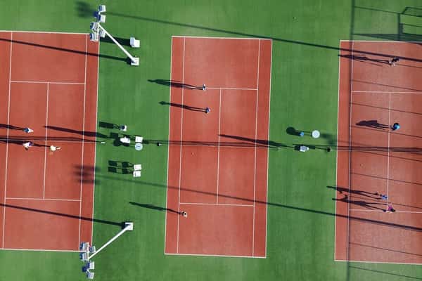 Aerial view of three clay tennis courts at Ikos Aria with players visibly engaged and shadows cast in the sunlight.