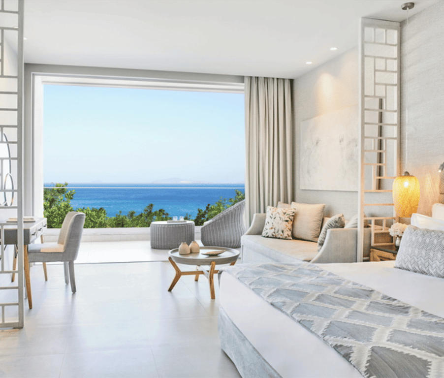 Ikos Aria junior suite with balcony view of the sea. Room includes modern decor, seating area, and outdoor balcony chairs.