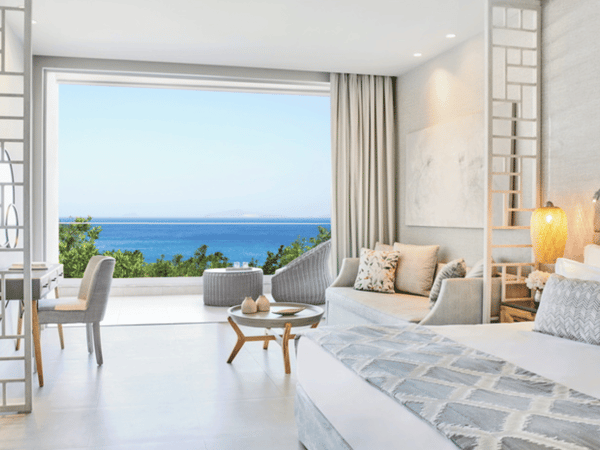 Ikos Aria junior suite with balcony view of the sea. Room includes modern decor, seating area, and outdoor balcony chairs.