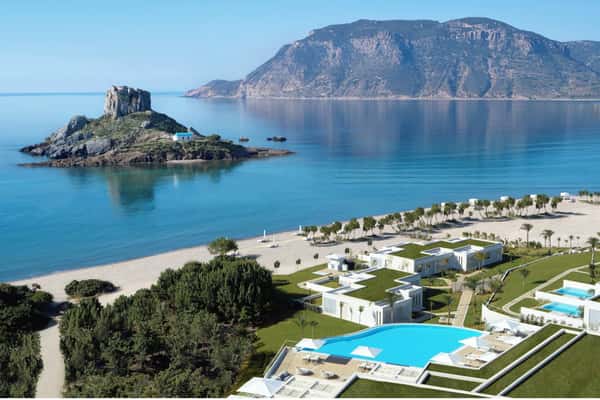 Aerial view of Ikos Aria resort in Kos, Greece, with contemporary white buildings, a pool, greenery, and the Aegean Sea.