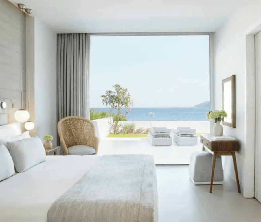 Sea view one-bedroom suite at Ikos Aria with private garden, featuring a bed, wicker chair, and large window overlooking the Aegean Sea.